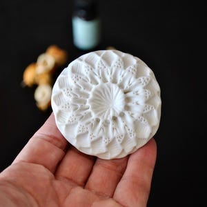 May include: A white, circular ceramic essential oil diffuser with a detailed floral pattern. The diffuser is held in a hand against a black background. A small bottle and dried flowers are in the background.