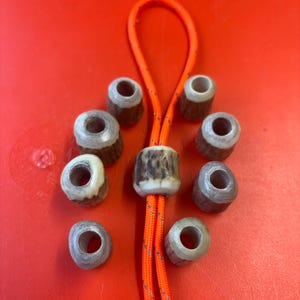 May include: A collection of eight cylindrical beads made from textured brown and white antler material, arranged around a central bead threaded onto a bright orange paracord loop with reflective silver stitching details.