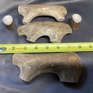 May include: A collection of natural antler dog chews. The chews are various sizes and shapes, with a light brown color and a textured surface. A yellow tape measure shows the length of the largest chew is approximately 9 inches.