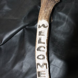 May include: A carved antler with the word "WELCOME" painted in black on a white background. The antler is a natural brown color and has a rough texture. The background is a dark, textured surface.