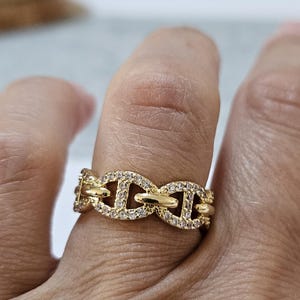 Könnte beinhalten: Ein goldfarbener Ring mit einem sich wiederholenden Ankerketten-Glieddesign. Jedes ovale Glied ist mit kleinen, klaren, funkelnden Steinen besetzt, die die gesamte Oberfläche des Metalls bedecken und ein durchgehendes, strukturiertes Band bilden.