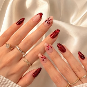 May include: A close-up of a hand with a manicure featuring long, oval-shaped nails. Some nails are painted a deep red with glitter, while others have a nude base with red bow designs. Gold rings adorn the fingers.