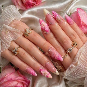 May include: Close-up of a hand with long, almond-shaped nails. The nails feature a pink and white gradient, 3D flower designs, rhinestones, and gold accents. The hand also wears gold rings with floral and gemstone details.