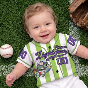 Może przedstawiać: Niemowlę w biało-zielonej koszulce baseballowej w paski z napisem "Buzz Lightyear" i numerem "00" w kolorze fioletowym. Na zdjęciu znajduje się r&oacute;wnież piłka do baseballu i rękawica do baseballu.