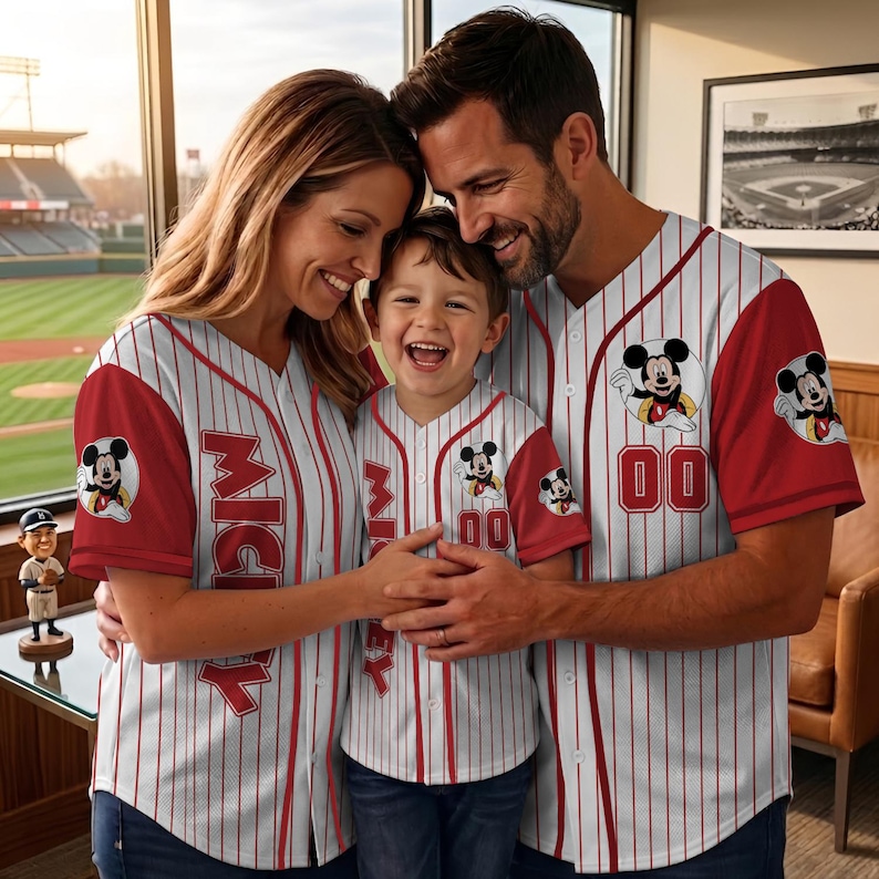 May include: Three people wearing matching baseball jerseys. The jerseys are white with red pinstripes and sleeves. The jerseys have a Mickey Mouse graphic on the sleeve and the word "Mickey" on the front. The jerseys also have the number "00" on the front.
