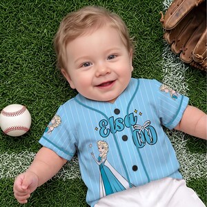 May include: A baby wearing a light blue baseball jersey with the name "Elsa" and a cartoon image of Elsa from Frozen. The jersey has white pinstripes and black buttons. A baseball and glove are in the background.