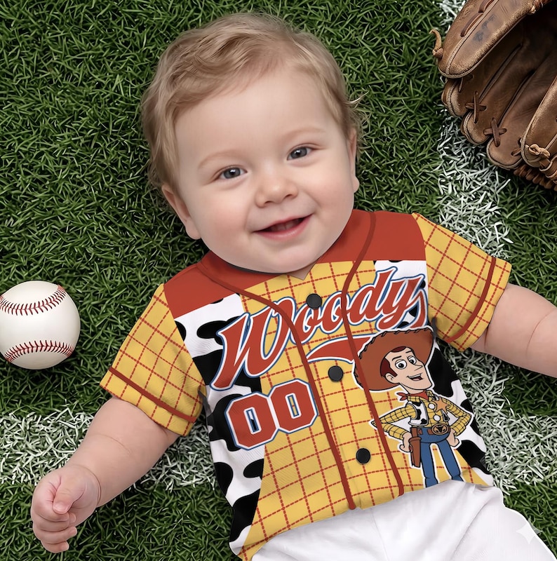 Puede incluir: Una camiseta de b&eacute;isbol de manga corta con botones, con un patr&oacute;n de cuadros amarillos, detalles de estampado de vaca en blanco y negro, y un gr&aacute;fico del personaje Woody. El frente muestra el texto Woody's y el n&uacute;mero 00 en letras rojas y azules.