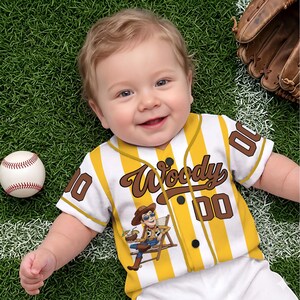 May include: A baby wearing a yellow and white striped baseball jersey with the name "Woody" and the number "00" on it. A baseball and a baseball glove are also in the image. The baby is lying on green grass.