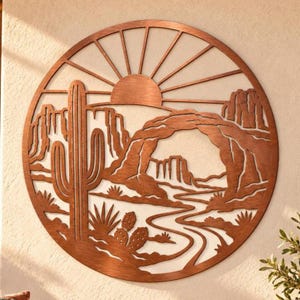 May include: A circular copper-toned metal wall art piece featuring a desert landscape. The design includes a tall saguaro cactus, smaller cacti, a winding path, rock formations, a natural stone arch, and a sunburst pattern at the top.