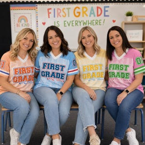 May include: Four v-neck jerseys in peach, light blue, yellow, and pink, each featuring vertical stripes, the text "FIRST GRADE" in bold letters, and "Mrs. Olivia" in script. Each sleeve displays the number "01" with two horizontal stripes.