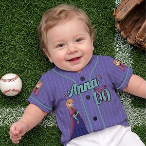 May include: A baby wearing a purple baseball jersey with the name "Anna" in teal script. The jersey has vertical pinstripes, black buttons, and images of a cartoon character. A baseball and a baseball glove are also in the image.