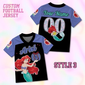 Puede incluir: Camiseta de f&uacute;tbol personalizada con una parte delantera negra, un dibujo animado de Ariel y el nombre "Ariel" en escritura blanca. La camiseta tiene mangas azul claro y cuello en V. La parte trasera es negra con "Your Name" y el n&uacute;mero "00".