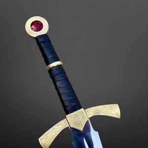 May include: A sword featuring a black leather-wrapped grip, a gold-toned crossguard with intricate etched patterns, and a circular pommel set with a faceted red gemstone. The blade is metallic silver with a central fuller running down the length.