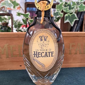 Peut inclure: Une bouteille en verre décorative avec un bouchon noir et un croissant de lune doré. La bouteille porte une étiquette avec le texte "LA MAGIE D' HECATE" et est ornée de deux ornements de serpent noirs. La bouteille est remplie d'une substance brune.