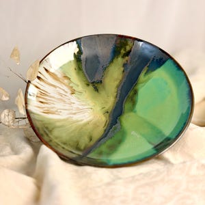 Könnte beinhalten: Eine runde Keramikschale mit einer glänzenden Glasur in den Farben Dunkelmarineblau, leuchtendes Smaragdgrün und Cremeweiß. Die Oberfläche zeigt ein fließendes, marmoriertes Muster mit dunklen Streifen, die von der Mitte zum Rand verlaufen.