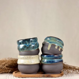 Peut inclure: Quatre petites tasses en céramique avec des bases gris foncé mates et des bords émaillés dans des tons bleu, vert et crème. Les tasses sont empilées par deux sur un petit plateau en bois, avec des glaçures coulantes sur les parties supérieures.