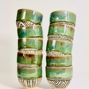 Op de afbeelding: Twee verticale stapels van elk vijf keramische bekers. De bekers hebben een glanzend groen glazuur aan de bovenzijde en een crèmekleurige basis met handgeschilderde zwarte geometrische patronen, waaronder lijnen, stippen en chevronvormen.