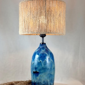 Puede incluir: Una lámpara de mesa de cerámica azul con una pantalla texturizada envuelta en cuerda. La base tiene un efecto de esmalte brillante y goteado. La lámpara es un elemento de iluminación decorativo.
