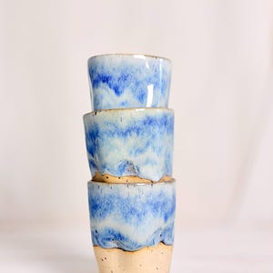 Könnte beinhalten: Ein Stapel aus drei Keramikbechern mit gesprenkelter beiger Basis und einer glänzenden blau-weißen Tropfglasur. Die Becher haben eine zylindrische Form mit leicht verjüngtem Boden und zeigen einen Übergang zwischen Ton und Glasur.