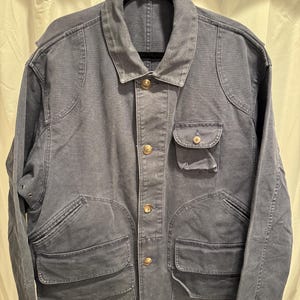 May include: A blue-grey jacket with a collared neckline and multiple pockets. The jacket has gold-colored buttons and a classic design. The jacket is hanging on a black hanger.
