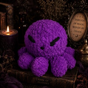 May include: A plush, purple octopus with black embroidered eyes and a mouth sits atop a vintage book. The octopus is made of a soft, textured material. The book reads "BOOK OF SHADOWS". A candle and other occult items are in the background.