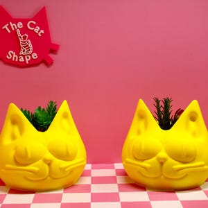 Op de afbeelding: Twee felgele, katvormige plantenbakken met gladde oppervlakken en gevormde gelaatstrekken, elk met een kleine groene plant. Een roze bord in de vorm van een kattenkop met de tekst The Cat Shape bevindt zich aan de muur boven de plantenbakken.