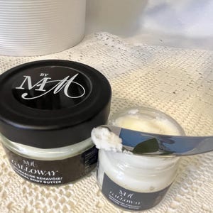 May include: Two small glass jars of body butter with black lids. One jar is open with a small amount of product on a silver knife. The jars have a label that reads "Galloway Natural Body Butter" and the initials "MM".
