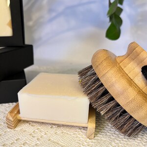 May include: A rectangular off-white bar of soap rests on a light-coloured wooden soap dish. Beside the soap dish is a round wooden body brush featuring a light bamboo handle and dense, dark brown bristles with a black fabric strap attached.