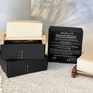 Op de afbeelding: Twee rechthoekige blokken talkzeep in gebroken wit op houten zeepbakjes, vergezeld van twee zwarte kartonnen doosjes. Eén doosje toont de tekst HIGHLAND TALLOW SOAP WITH SIBERIAN FIR, ingrediënten, waarschuwingen en een nettogewicht van 75g.