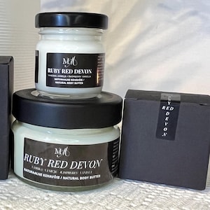 Op de afbeelding: Twee glazen potten body butter met zwarte deksels zijn op elkaar gestapeld. De potten hebben etiketten met de tekst "RUBY RED DEVON" en "NATURAL BODY BUTTER". Een kleine zwarte doos is ook aanwezig, met de woorden "RED DEVON" verticaal afgedrukt.