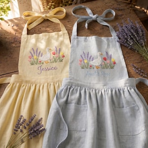 Op de afbeelding: Twee schorten, een gele en een lichtblauwe, met geborduurde ontwerpen. Elk schort heeft een eend, bloemen en een naam. Het gele schort zegt "Jessica" en het blauwe schort zegt "Amelia Rose". Beide schorten hebben een strik bij de nek.