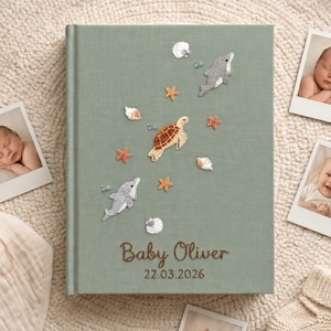 May include: Sage green baby photo album with embroidered sea creatures, including dolphins, a turtle, and starfish. The album is personalized with the name "Baby Oliver" and the date 22.03.2026.