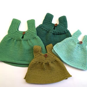 May include: Five hand-knitted dresses in various shades of green and olive. Each dress has a scoop neckline, shoulder straps, and a wooden button fastening. The dresses are displayed on a white background, highlighting the different green hues.