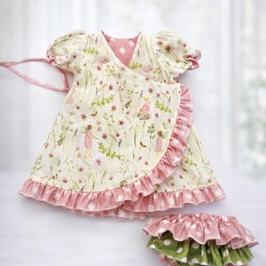 May include: A cream-colored baby dress and diaper cover set. The dress features a wrap design with a pink polka dot lining and ruffle trim. The fabric is printed with a floral and fairy design.