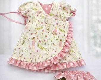 May include: A cream-colored baby dress and diaper cover set. The dress features a wrap design with a pink polka dot lining and ruffle trim. The fabric is printed with a floral and fairy design.
