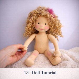 May include: A handmade doll with a light skin tone, curly blonde hair, and a pink bow. The doll is approximately 13 inches tall. The doll has rosy cheeks and is sitting on a white, textured surface. The text "13" Doll Tutorial" is at the bottom.