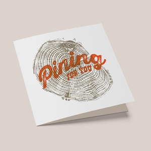 Puede incluir: Tarjeta de felicitación blanca plegada con un diseño de anillos de árbol en marrón y gris. Las palabras "Pining for You" están impresas en naranja. La tarjeta está en blanco por dentro, adecuada para varias ocasiones.