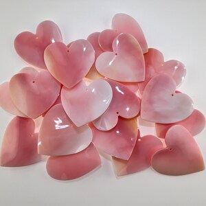 Puede incluir: Una colección de colgantes en forma de corazón hechos de material de concha rosa suave e iridiscente. Cada corazón individual presenta un pequeño orificio circular cerca del centro superior, lo que permite una fácil fijación a joyas o cordones.