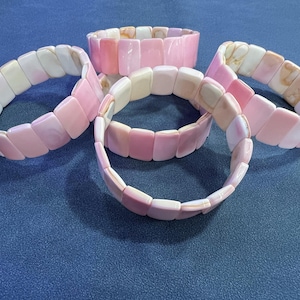 May include: Five stretch bracelets with rectangular, pink and white beads. The bracelets are arranged on a blue surface. The beads have a polished, glossy finish. The bracelets are a mix of light and dark pink.