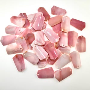 May include: A collection of numerous flat, pentagonal-shaped pink shell pendants. Each piece features a smooth, polished surface with natural white and light pink marbled variations and a single small hole drilled near the top edge for hanging.