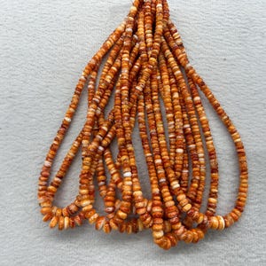 May include: A collection of orange and white beaded necklaces. The beads are small and uniform in size, strung together to create multiple strands. The necklaces are arranged in a cascading pattern against a light gray background.