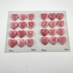 Puede incluir: Veinticuatro cabujones en forma de corazón en tonos rosa y blanco, dispuestos en dos grupos de doce sobre tarjetas blancas. Cada tarjeta está etiquetada como 20mm y 12pcs. Los corazones lisos y brillantes presentan patrones de color concéntricos.
