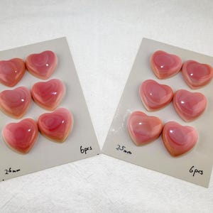 May include: Two white display cards each holding six pink heart-shaped cabochons. Each card features the handwritten text 25mm and 6pcs. The hearts have a smooth, glossy finish with layered pink and white tones throughout the surface.