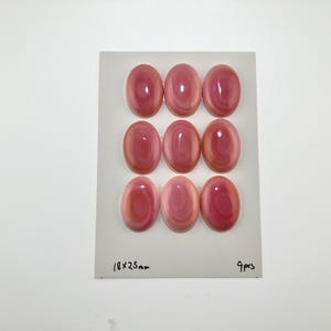Puede incluir: Nueve cabujones ovalados de color rosa con un acabado liso y brillante y sutiles patrones de remolinos, dispuestos en una cuadrícula de tres por tres sobre una tarjeta blanca. La tarjeta muestra el texto 18x25mm y 9pcs en la parte inferior.