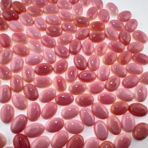 Puede incluir: Una colección de numerosos cabujones rosas pequeños, lisos y de forma ovalada. Cada pieza presenta una superficie pulida y brillante con tonos variados desde el rosa claro al rosa intenso, mostrando una forma abovedada uniforme.
