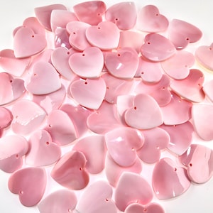 May include: A collection of numerous heart-shaped beads crafted from smooth, polished pink shell material. Each individual heart features a small, centered hole near the top, designed for threading. The pieces exhibit varying shades of soft pink.