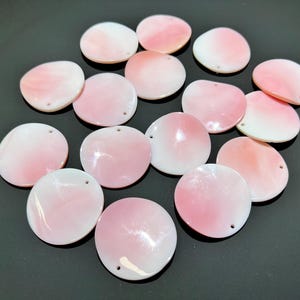 May include: A collection of round, flat shell beads featuring a smooth, iridescent finish with a gradient of soft pink and white colours. Each circular piece has a single small hole drilled near the edge for stringing, showcasing a natural lustre.