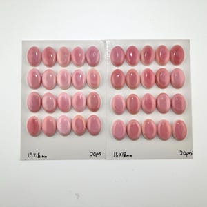 Puede incluir: Cuarenta cabujones ovalados de color rosa dispuestos en dos tarjetas de presentación rectangulares blancas. Cada tarjeta contiene veinte piezas, con el texto 13x18 mm y 20pcs impreso en la parte inferior. Los cabujones tienen una superficie lisa y pulida.