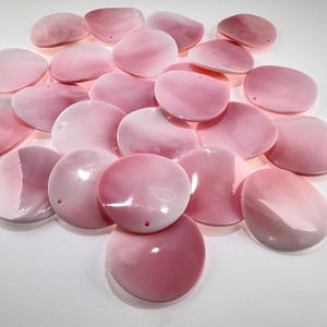 May include: A collection of round, flat, pink shell beads with a smooth, glossy finish. Each bead features a single small hole near the edge for stringing. The beads vary slightly in shape and size, displaying natural color gradients from light to dark pink.