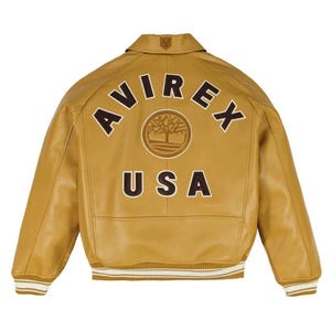 May include: A tan leather jacket featuring a classic collar and ribbed cuffs and hem with white stripes. The back displays the text AVIREX in a dark brown arch, a circular tree logo emblem in the center, and the text USA in dark brown letters below.